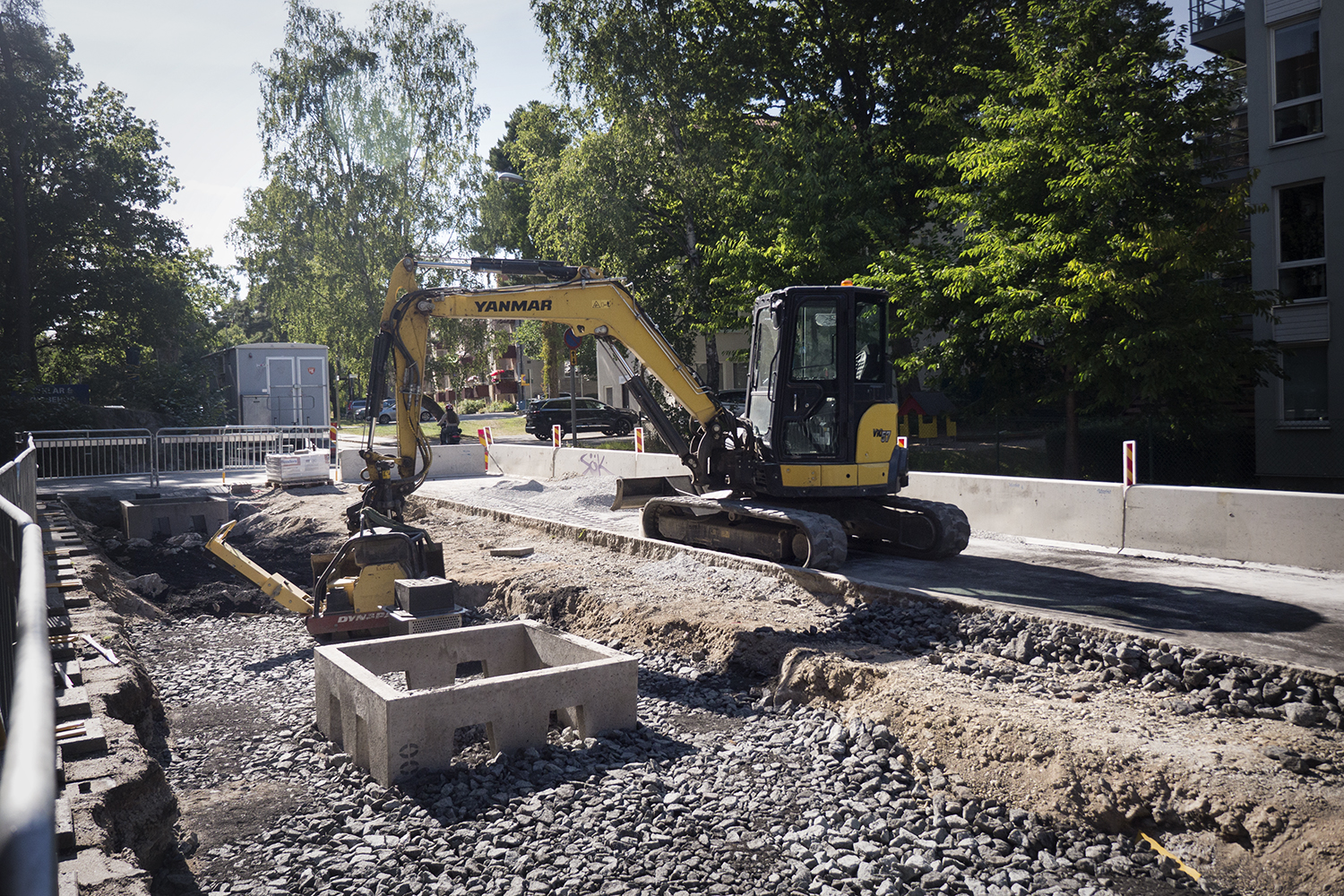 Kolmakadam En gul grävmaskin arbetar på en vägbyggnadsarbetsplats med grus- och betongkonstruktioner, omgiven av träd och byggnader i starkt solljus.